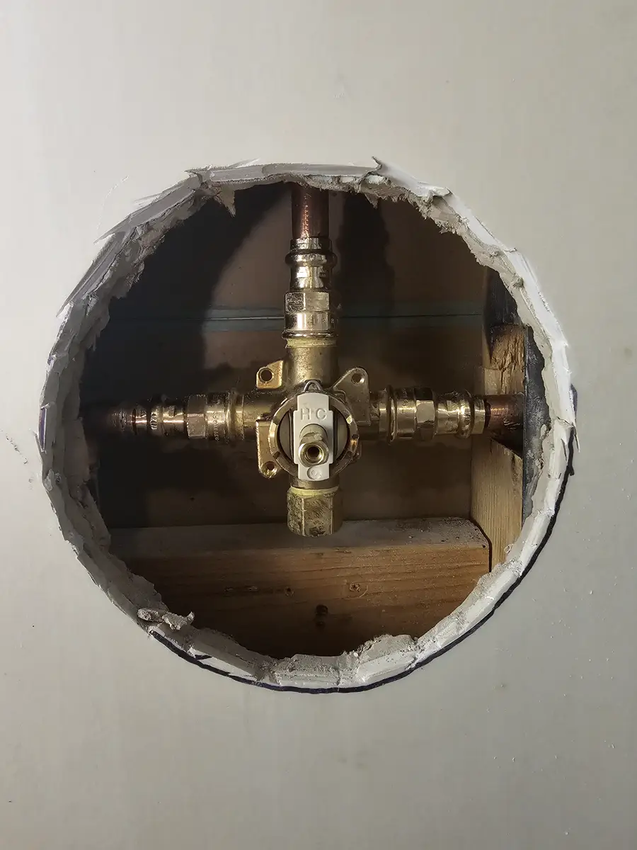 A brass shower valve with copper pipes, seen through a round hole cut in a drywall wall.