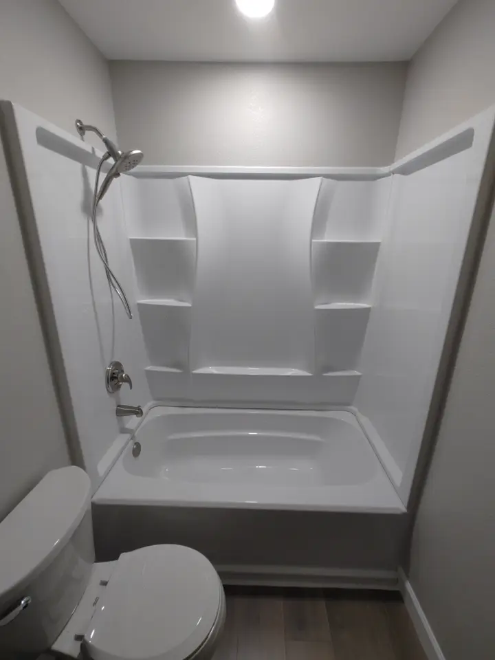 A white one-piece tub and shower unit with built-in shelves, a handheld shower head, and a chrome valve.