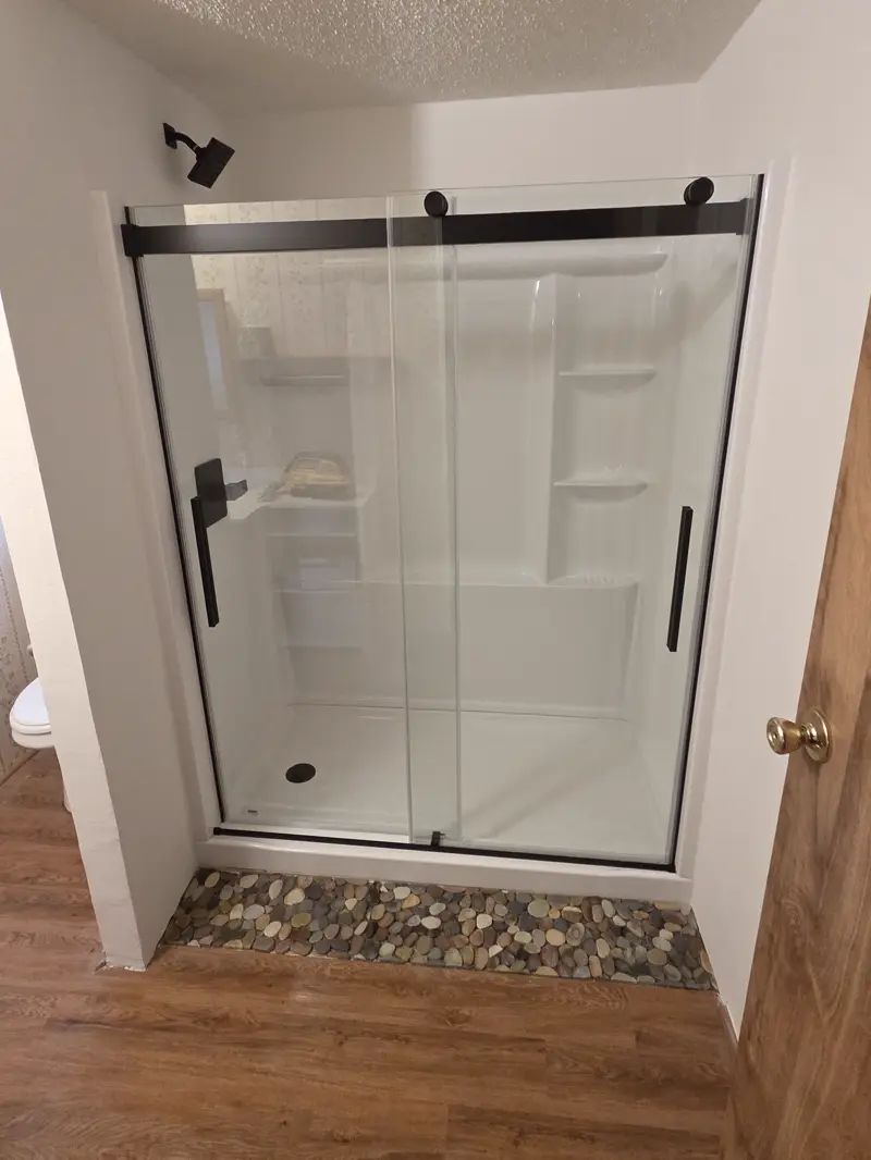 A shower with white wall panels and a sliding glass door with black hardware, and a strip of round pebbles on the floor in front.